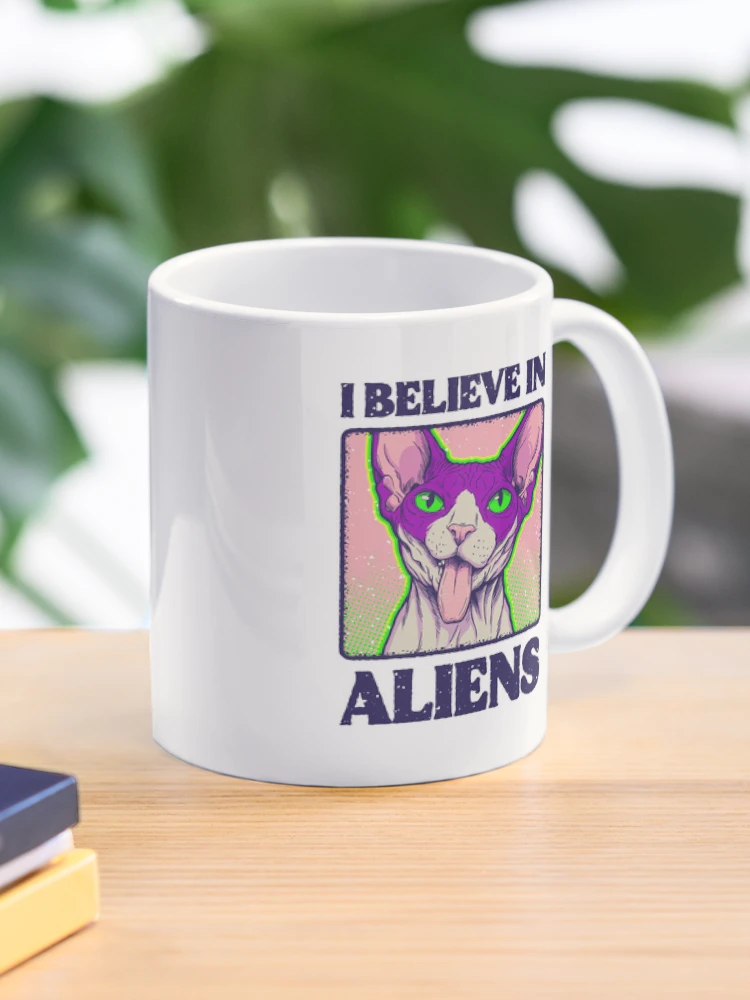 Ceramic white coffee mug featuring a colorful wrap-around 'I Believe in Aliens' cat design on a desk.